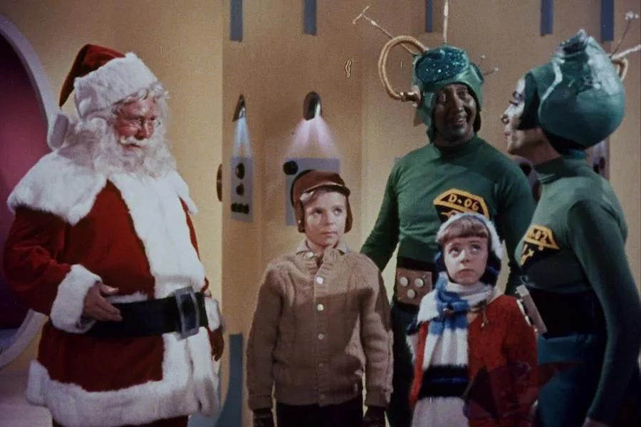 A still from a vintage sci-fi show shows Santa Claus standing beside two children and two alien characters in green…