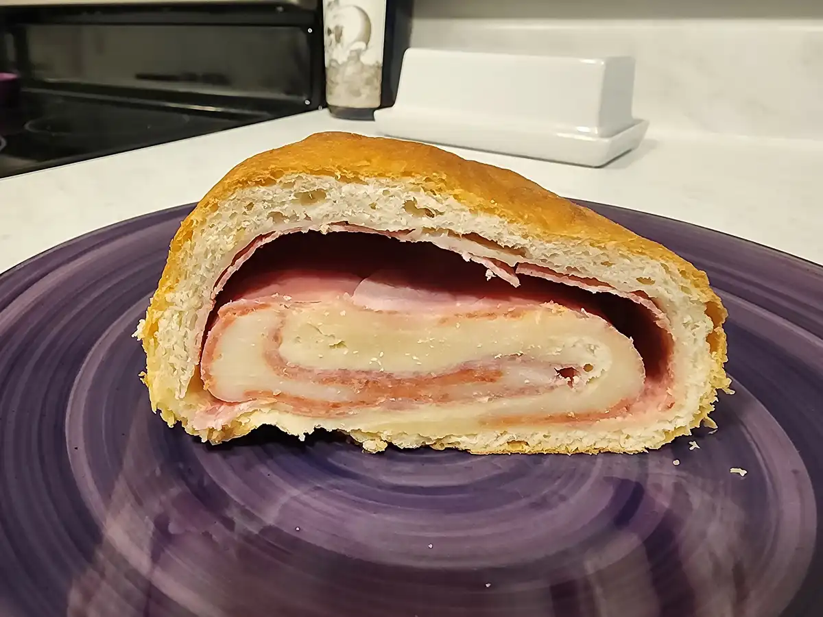 A sliced open bread roll reveals a spiral of ham and cheese inside, resting on a purple plate in a kitchen. The overlaid…