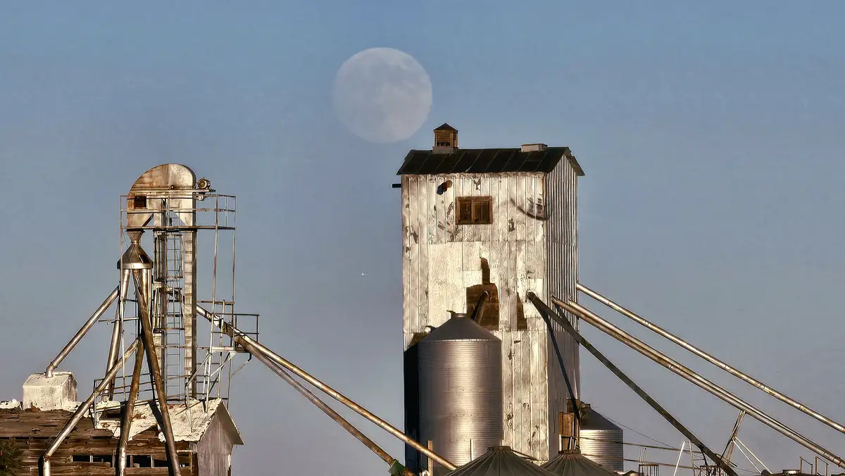 A photograph shows two weathered grain silos against a clear blue sky with a large, full moon visible. The moon is…