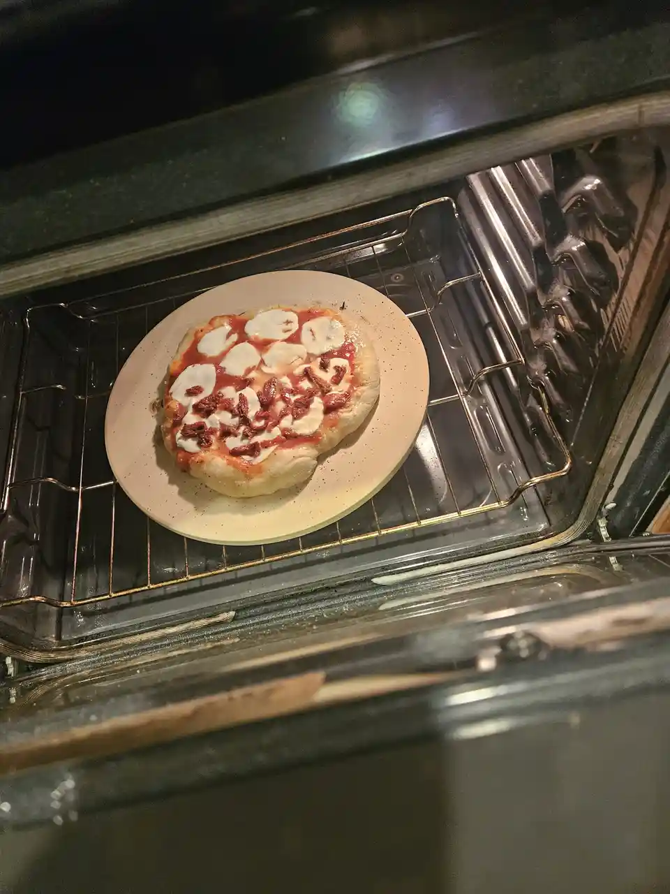 A homemade pizza with tomato sauce, mozzarella cheese, and sun-dried tomatoes sits on a light-colored plate inside a home…