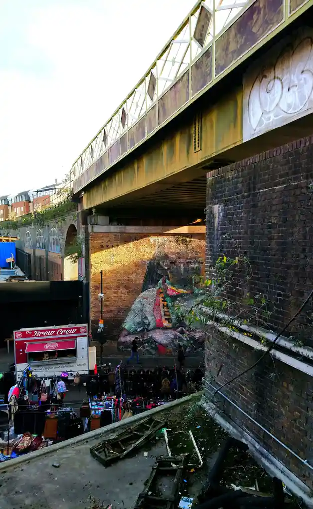 A photo of an urban underpass with a large mural of a person on a brick wall, a food truck labeled 'The Brew Crew', and…