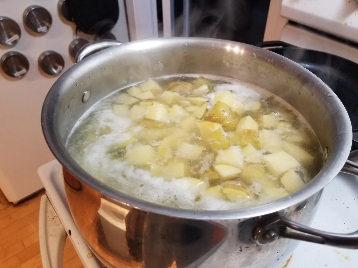 A stainless steel pot sits on a white stovetop, filled with vigorously boiling water and chopped potatoes, with steam rising…