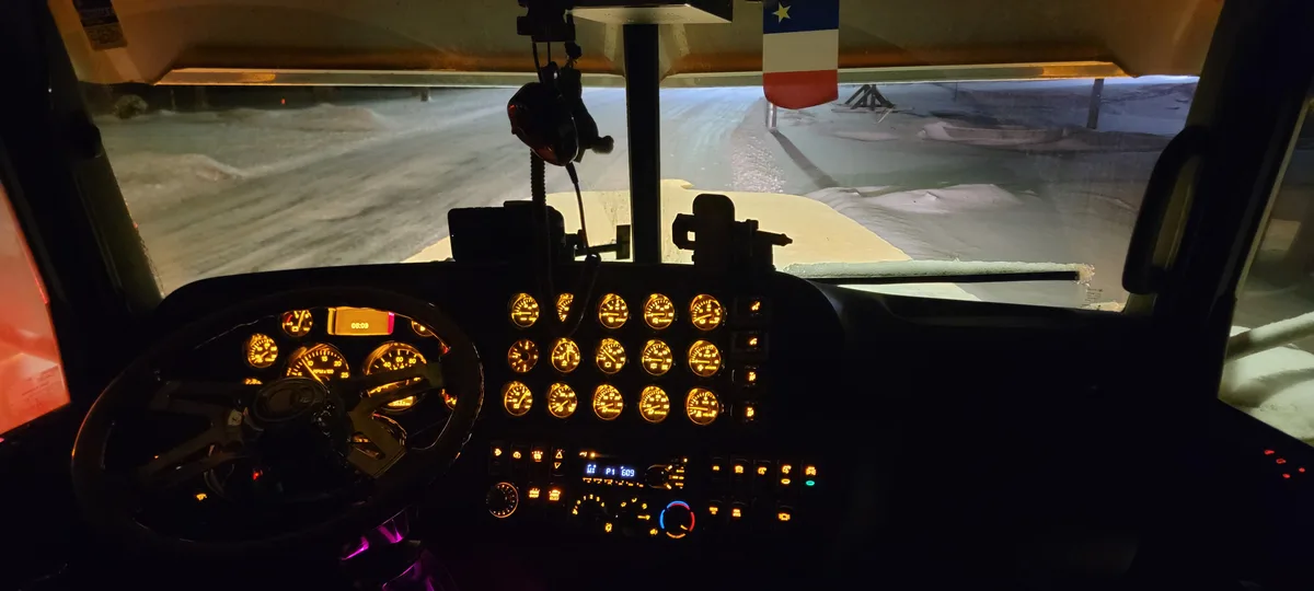 The image shows the interior of a snowplow truck's cab at night, with illuminated gauges and a steering wheel visible…