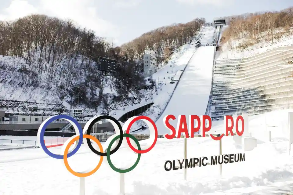 A snowy outdoor scene features the iconic Olympic rings in the foreground, with a large ski jump and tiered spectator stands…