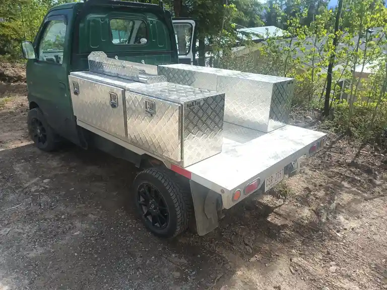 a green compact truck with a flatbed is parked on dirt, heavily modified with multiple large, silver diamond-plate aluminum…