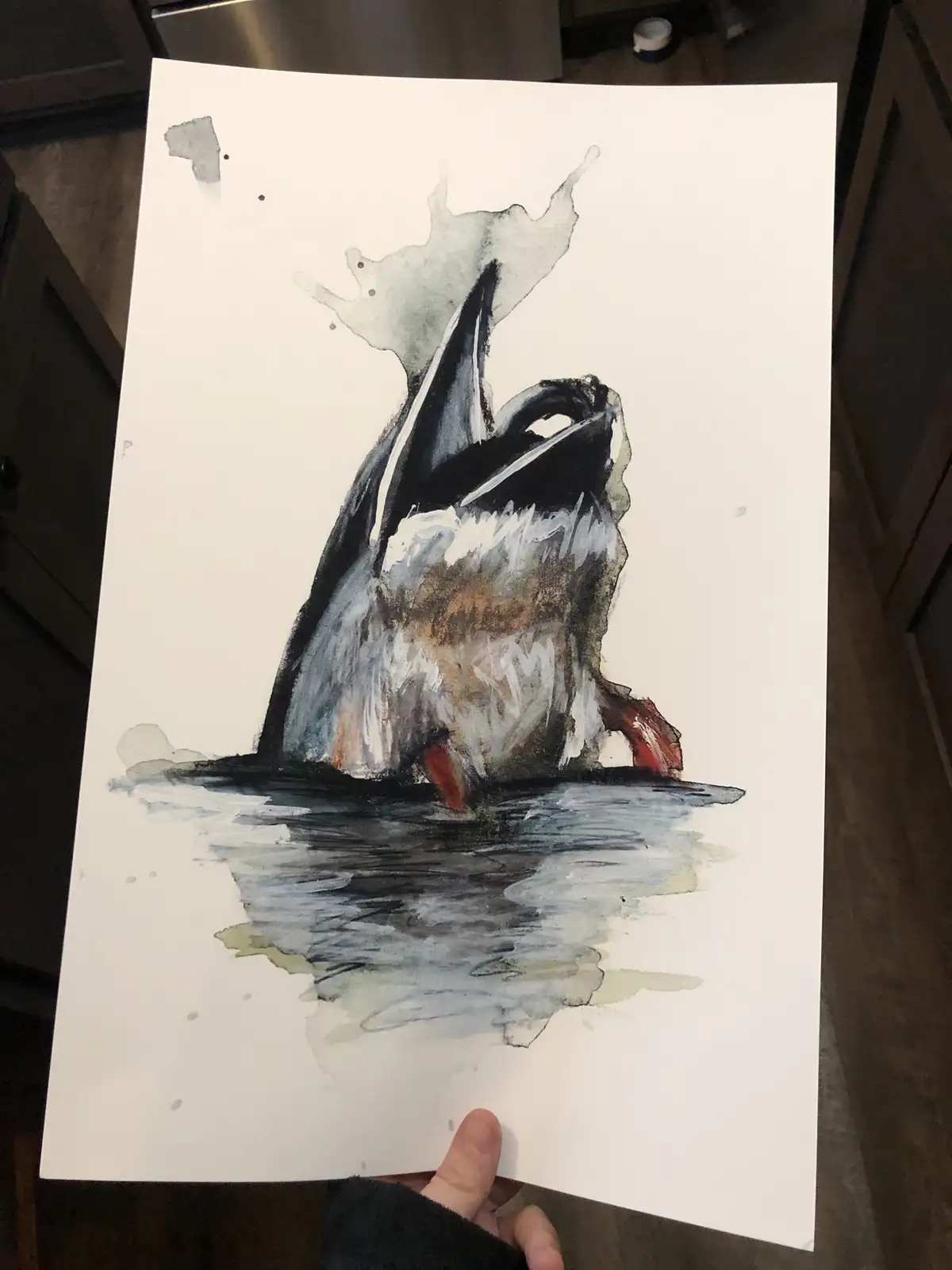 A person holds up a watercolor painting of a whale breaching out of water, with visible splatters and a somewhat abstract…