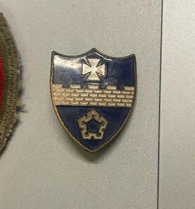 a close-up photo shows a shield-shaped military-style badge pinned to a gray surface, featuring a blue background with a…