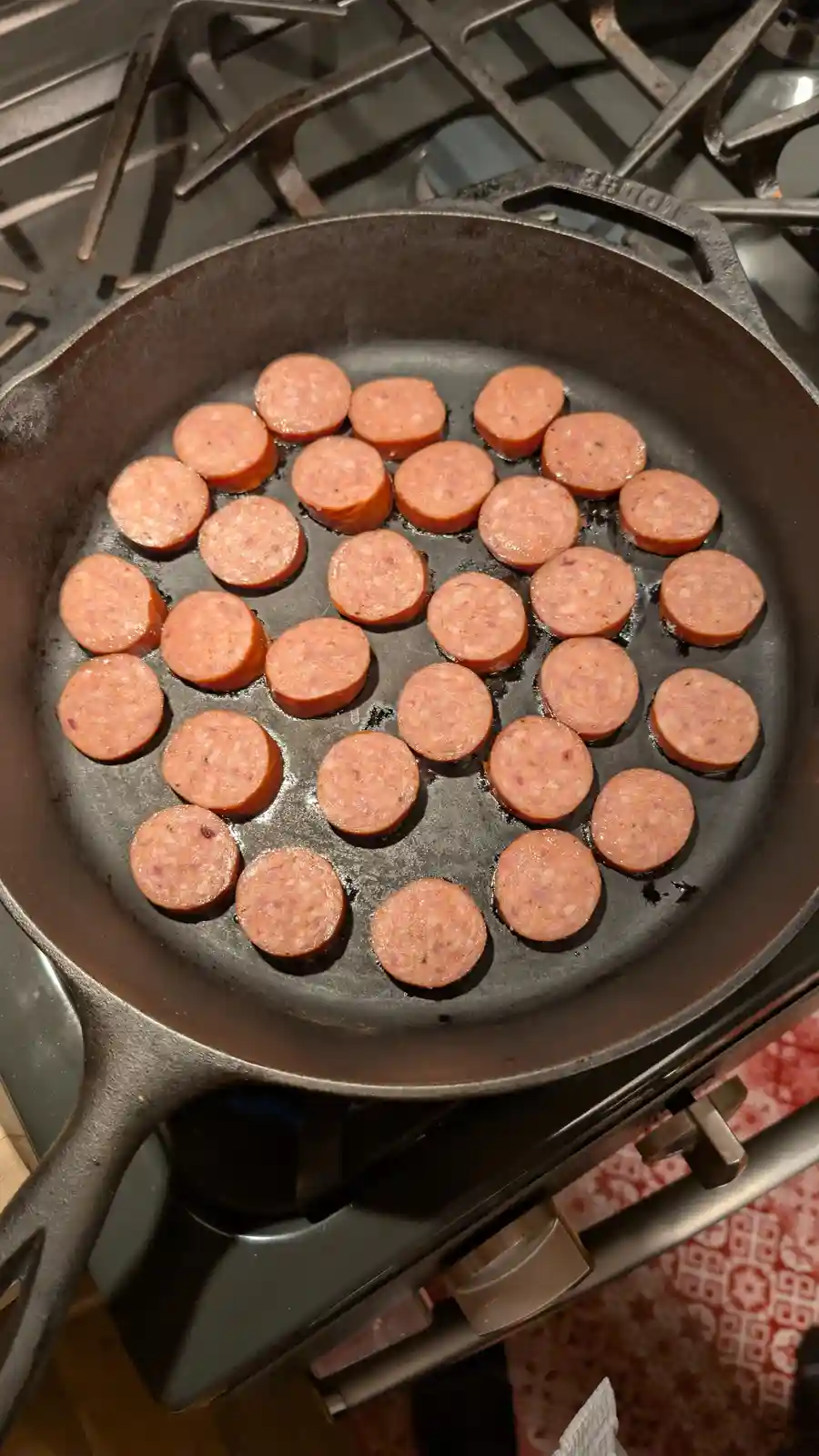A cast iron skillet on a gas stove contains numerous round slices of sausage cooking in oil. The text overlaid on the image…