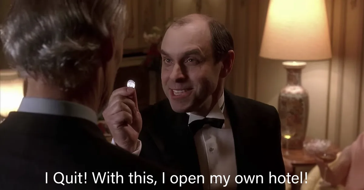 A man in a tuxedo holds up a small shiny object while smiling triumphantly at another man whose back is to the camera. The…
