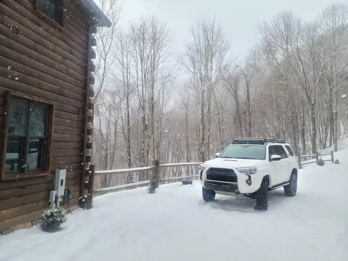A white Toyota 4Runner SUV is parked in deep snow beside a wooden log cabin during a snowstorm, with bare trees in the…