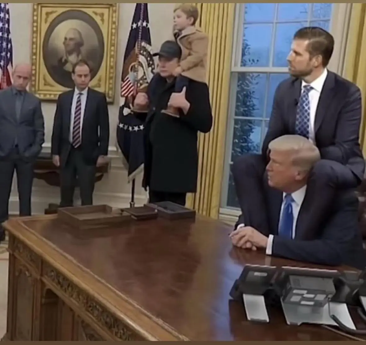 In the Oval Office, a man with a child on his shoulders gives a thumbs-up while standing near flags and a portrait of…