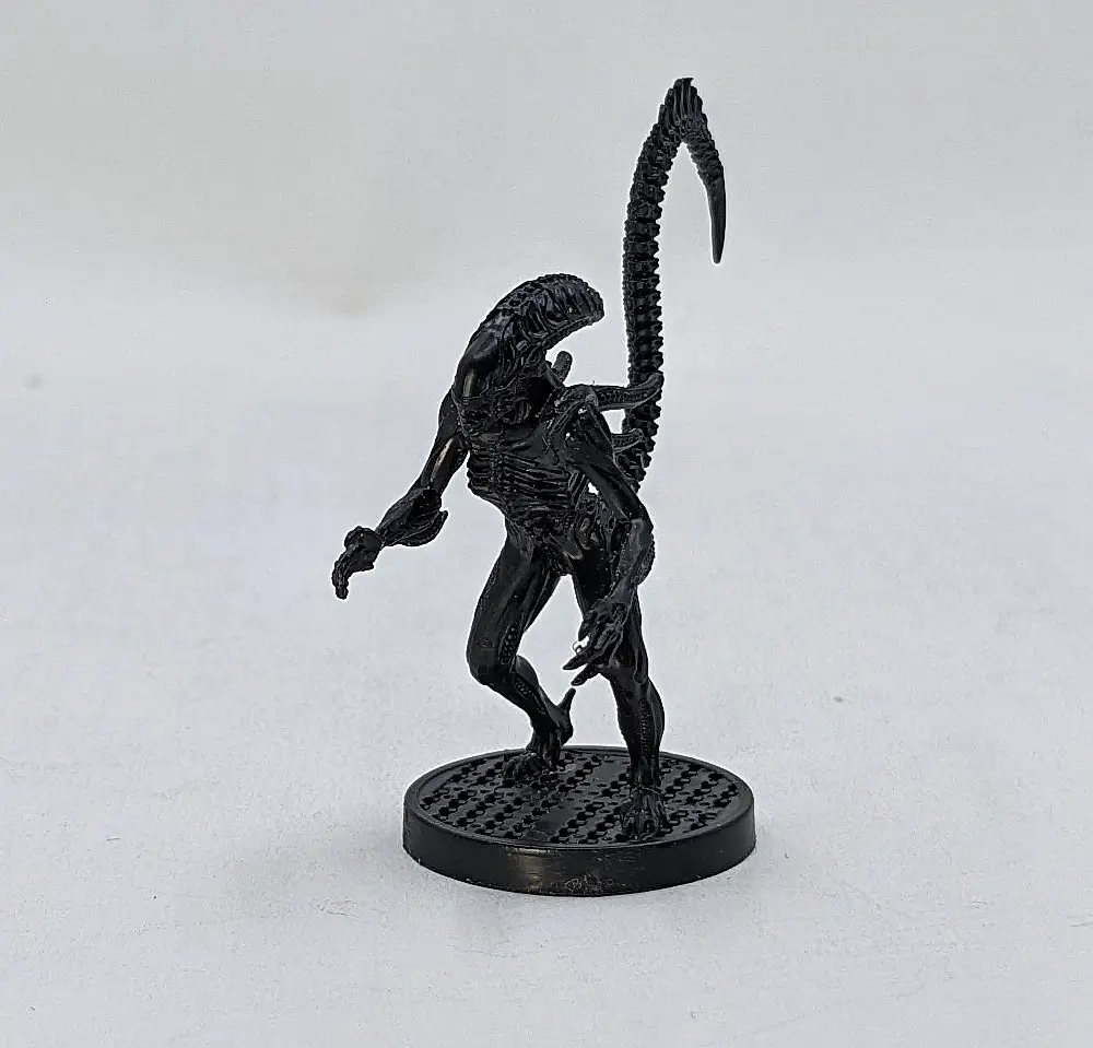 A black miniature figurine of the Xenomorph from the Alien franchise stands on a circular base against a plain white…