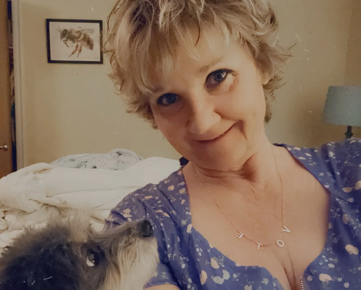 A woman with short blonde hair and a blue patterned top smiles at the camera while holding a small dog. She wears a necklace…