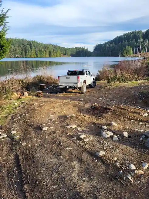 A white Hummer pickup truck is parked on a muddy, rocky dirt road next to a calm lake, surrounded by dense forest. Stacks of…