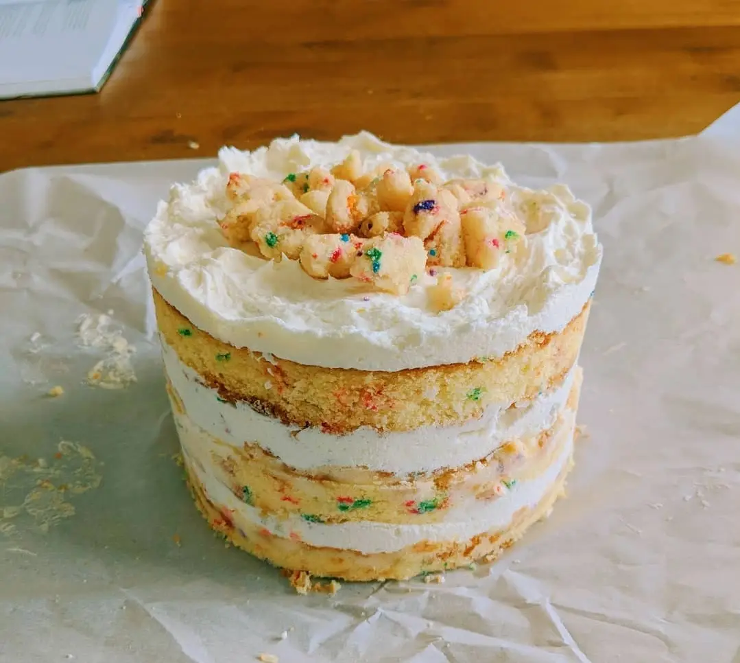 A multi-layered, unfrosted funfetti cake with white frosting and colorful sprinkles sits on crumpled parchment paper on a…