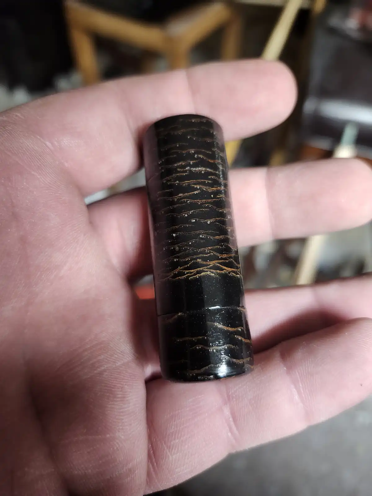 a hand holds a small, dark, cylindrical wooden object with wavy, golden-brown grain patterns resembling a mountain range…