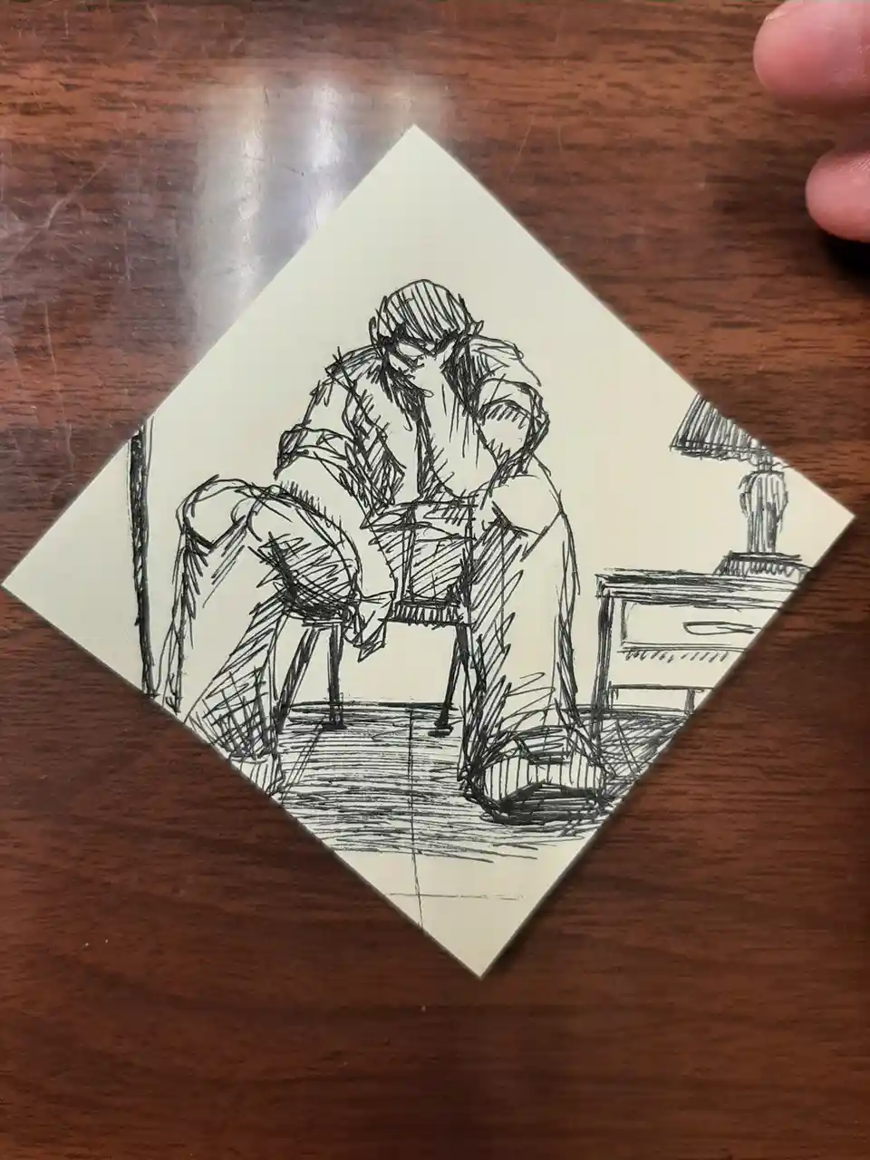 A hand-drawn sketch on a square piece of paper, rotated 45 degrees, depicts a person sitting at a desk with a lamp, hunched…