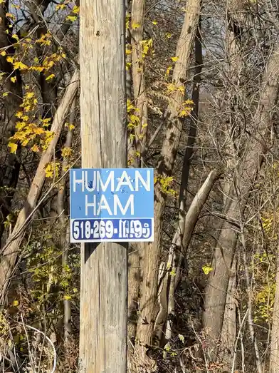 A blue sign with white text reading 'HUMAN HAM' and a phone number '518-269-1195' is affixed to a weathered wooden utility…
