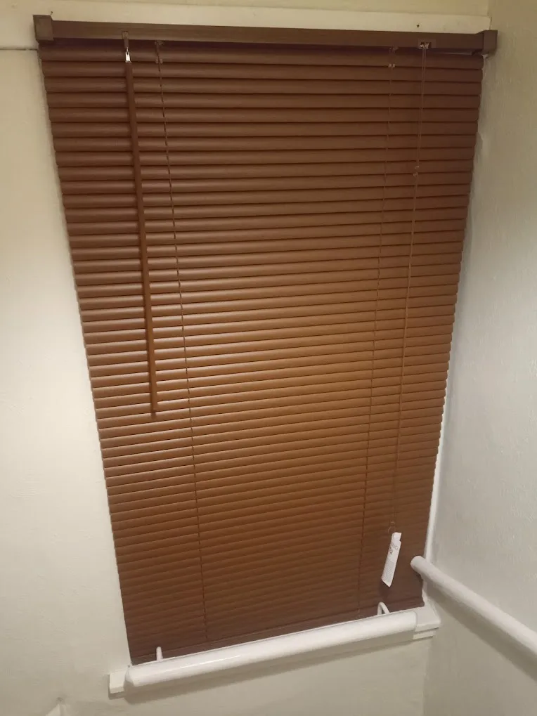 A close-up photo of brown horizontal window blinds covering a window, with a white plastic grab bar visible below and to the…