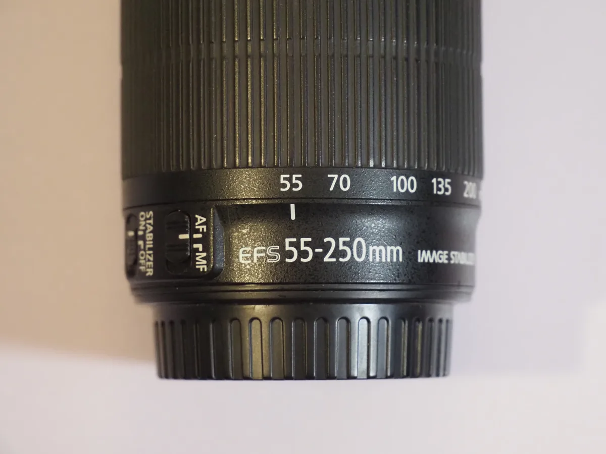 A close-up photo of a black Canon camera lens showing the text 'eFS 55-250mm' and focal length markers (55, 70, 100, 135…