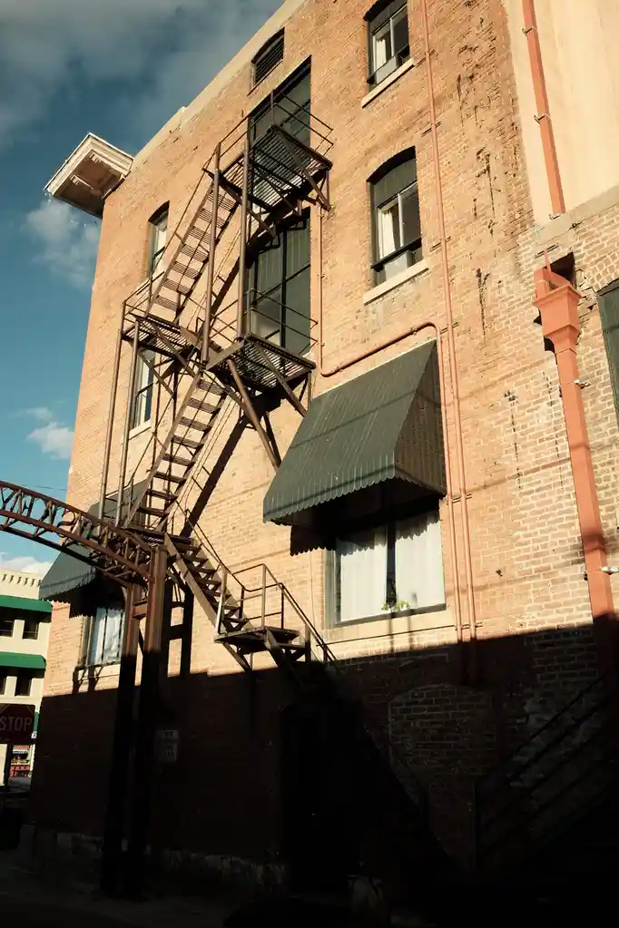 A low-angle photo shows a brick building with a prominent metal fire escape casting long shadows on its facade under a…