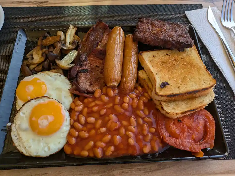 A full English breakfast is served on a black plate, featuring fried eggs, baked beans, sausages, bacon, mushrooms, toast…