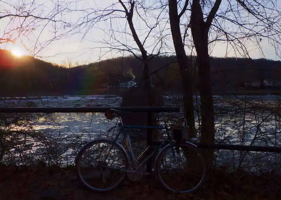 A teal bicycle is parked against a railing beside a partially frozen river at sunset, with bare trees silhouetted against…