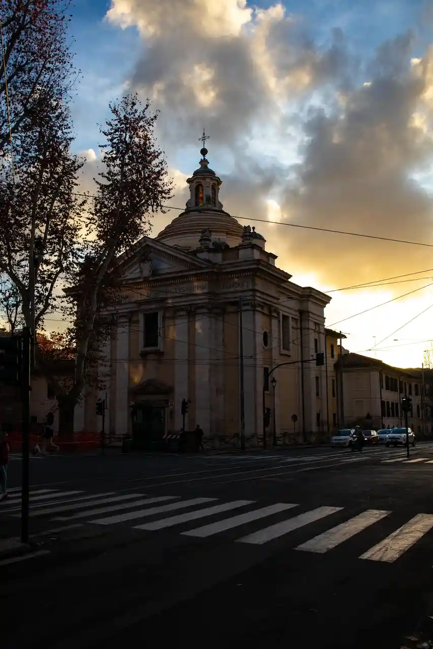 A photograph of a classical church with a dome and cross, set against a dramatic sunset sky with clouds. The foreground…