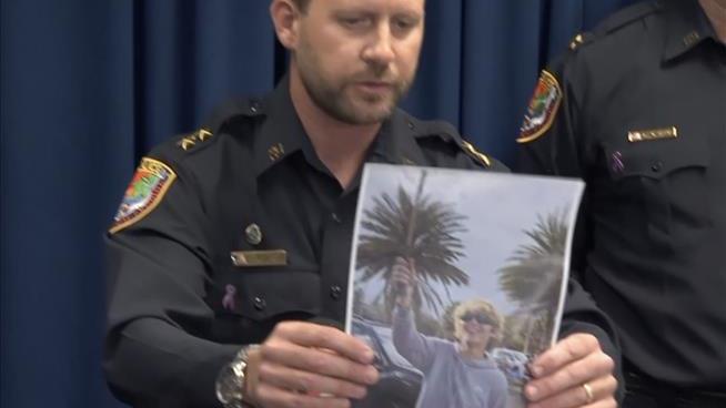 A police officer in uniform holds up a photo of a smiling woman with blonde hair and sunglasses, standing outdoors with palm…