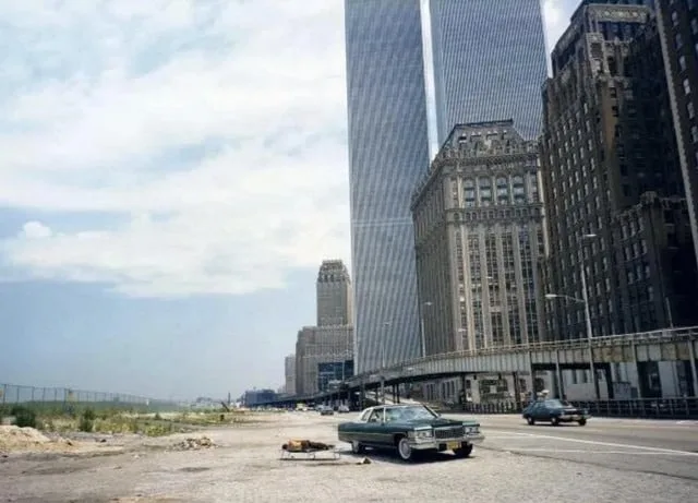 A vintage photo shows a green 1970s car parked on a dirt lot beside a road with a few other cars, in front of a cluster of…