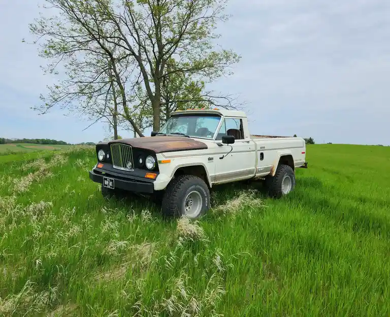 A white, rusty, lifted Jeep pickup truck with a brown hood sits parked in tall green grass under a tree. The overlaid text…