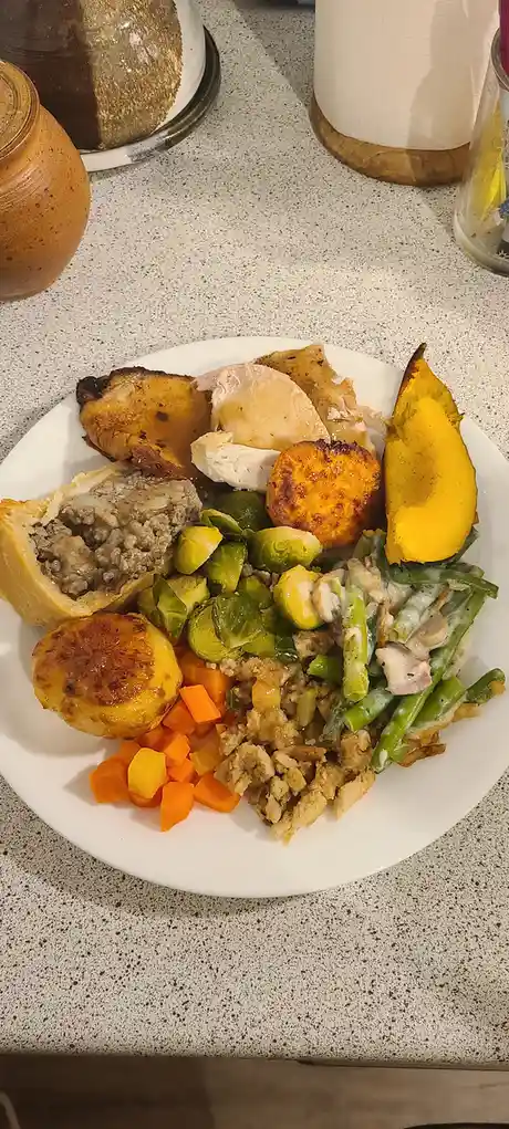 A white plate on a speckled countertop is piled high with an assortment of cooked foods including roasted chicken, sweet…