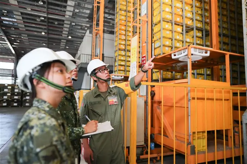 three people in military-style uniforms and white hard hats stand in a large warehouse with tall orange shelving units…