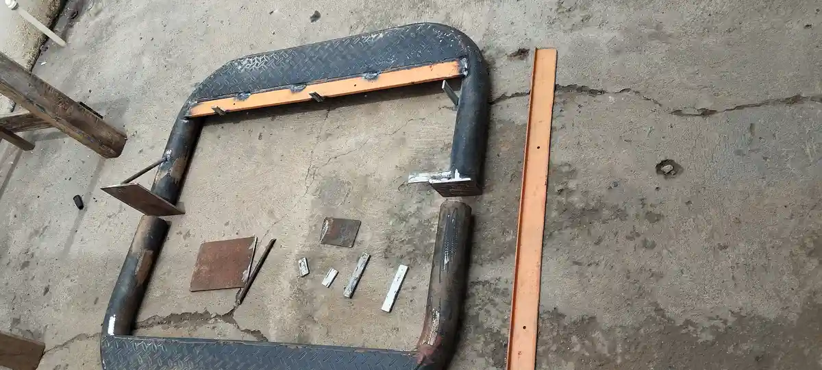 A partially assembled metal step stool lies on cracked concrete, constructed from a curved truck bumper with diamond plate…