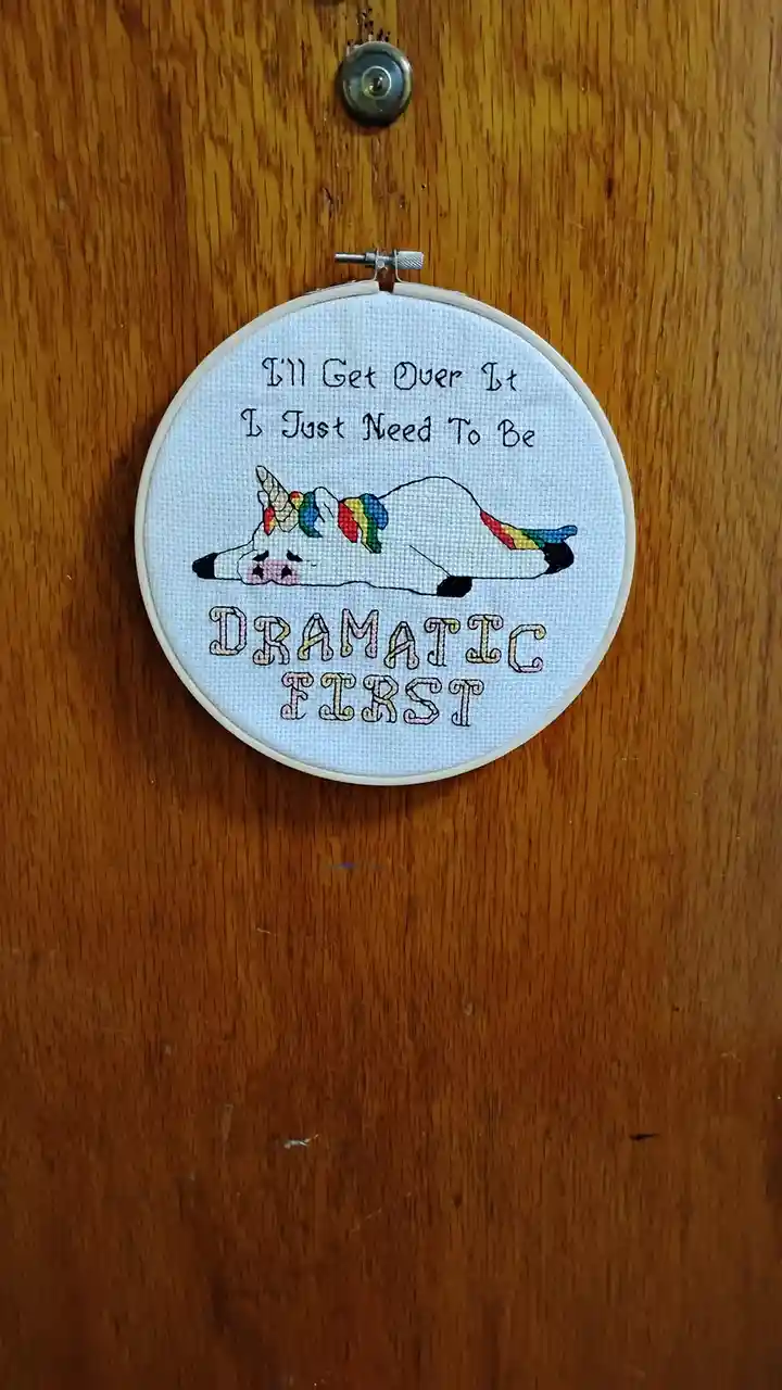 A cross-stitched embroidery hoop hangs on a wooden door, featuring a cartoon unicorn with a rainbow mane and tail lying…