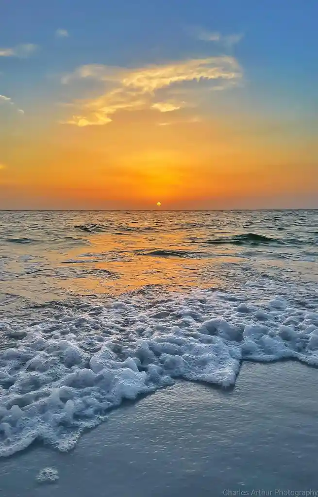 A serene sunset photograph shows the sun dipping below the horizon over a calm ocean, casting warm orange and yellow hues…
