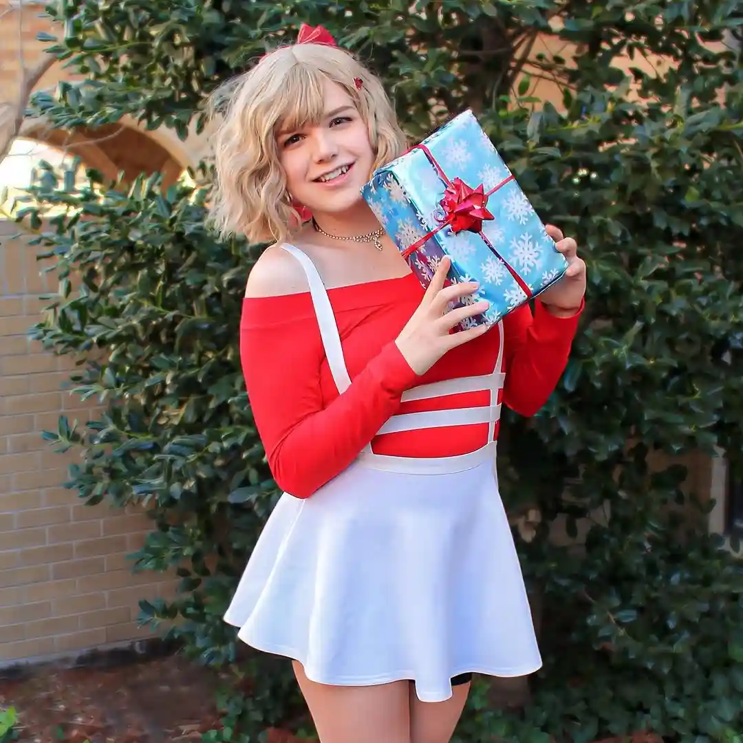 A person with blonde hair wearing a red and white outfit resembling a Santa helper costume holds a large blue gift wrapped…