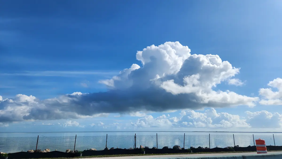 A scenic photo shows a large, dramatic white and gray cloud formation over a calm body of water, viewed through a chain-link…