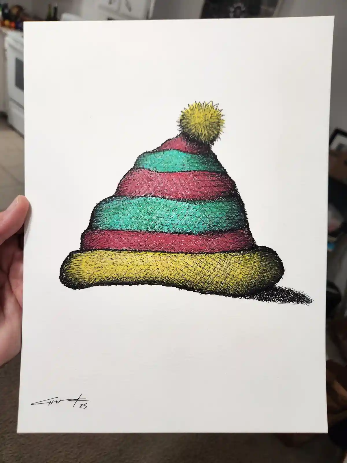 A hand holds a white paper displaying a colorful, hand-drawn illustration of a striped knit hat with a yellow pom-pom on…