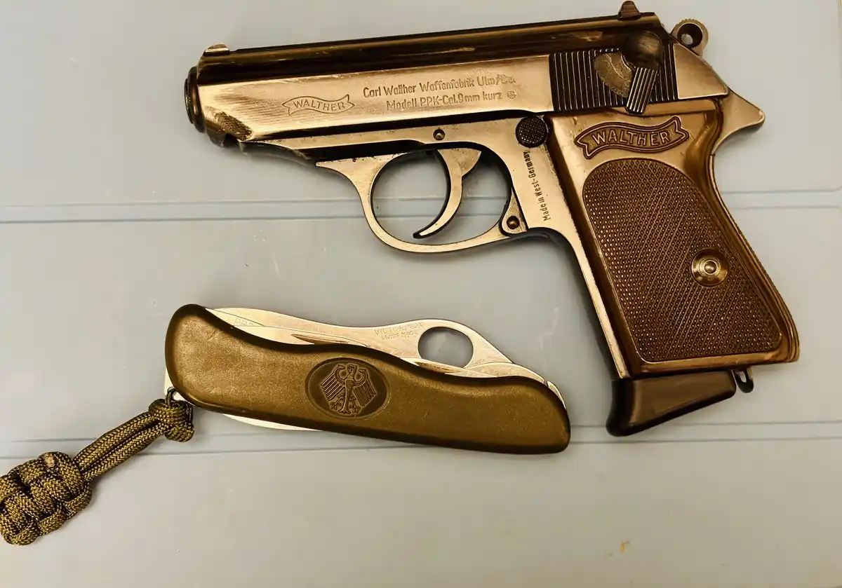 A gold-plated Walther PPK pistol is laid next to a gold-plated Victorinox Swiss Army knife, both with matching metallic…