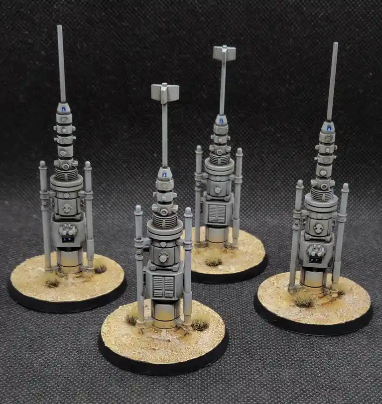 Four intricately painted miniature sci-fi towers with antennas and glowing blue lights stand on circular bases with desert…