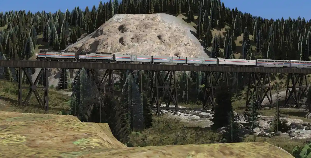 An Amtrak passenger train travels across a wooden trestle bridge through a forested mountain valley, with a large exposed…
