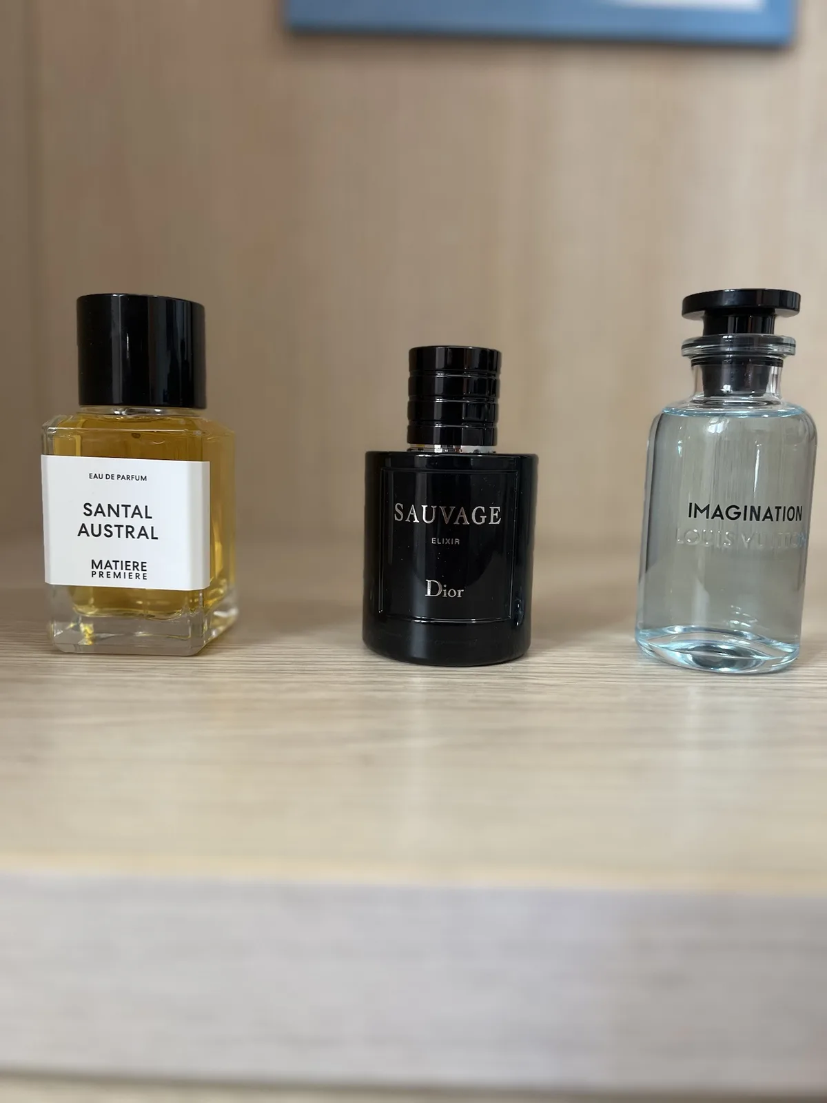 Three perfume bottles sit side by side on a wooden shelf: left is 'Santal Austral' by Matière Première with amber liquid…