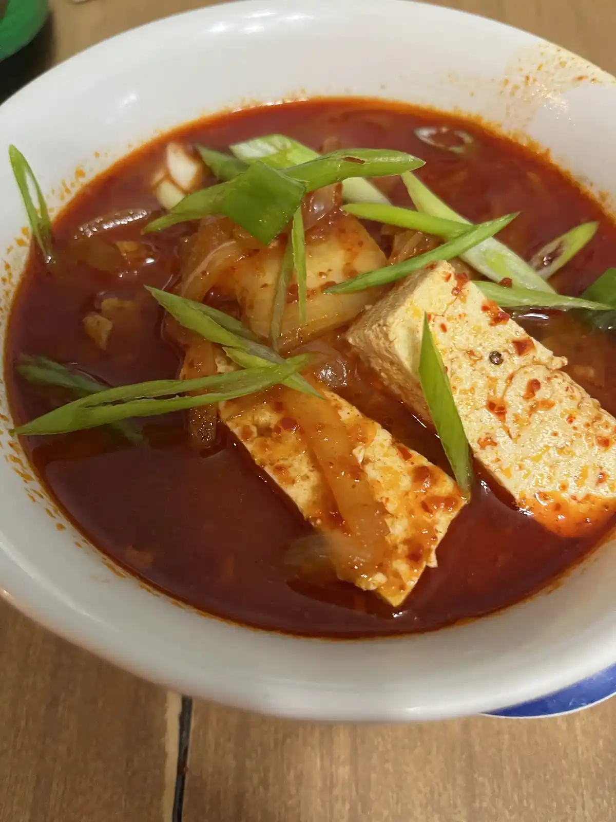 A close-up photo shows a white bowl filled with a thick, red kimchi stew, containing visible chunks of tofu and sliced green…