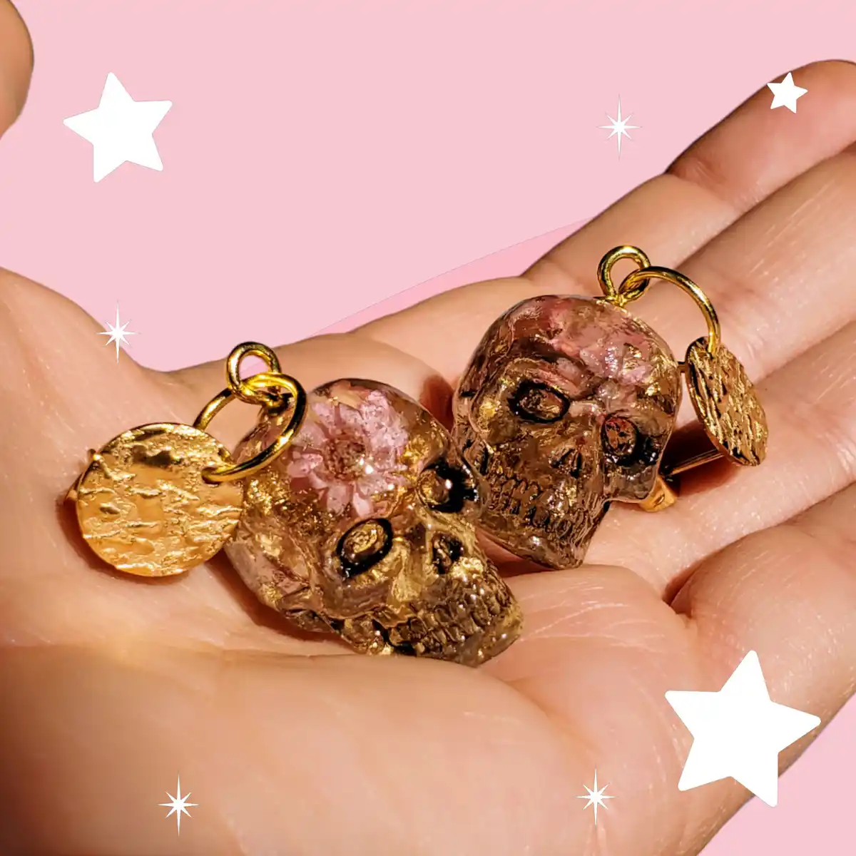 A hand holds two small skull-shaped charms with pink crystal interiors and gold accents against a pink background with white…