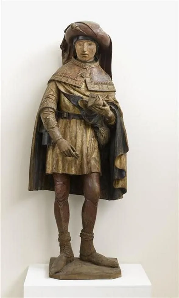 A wooden statue of a man in historical European clothing, including a hat, cape, and boots, stands on a white pedestal. The…