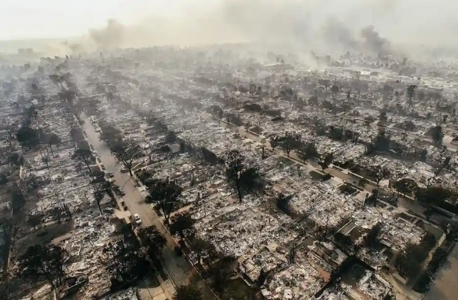 An aerial view of a residential neighborhood completely destroyed by fire, with charred rubble, blackened trees, and smoke…