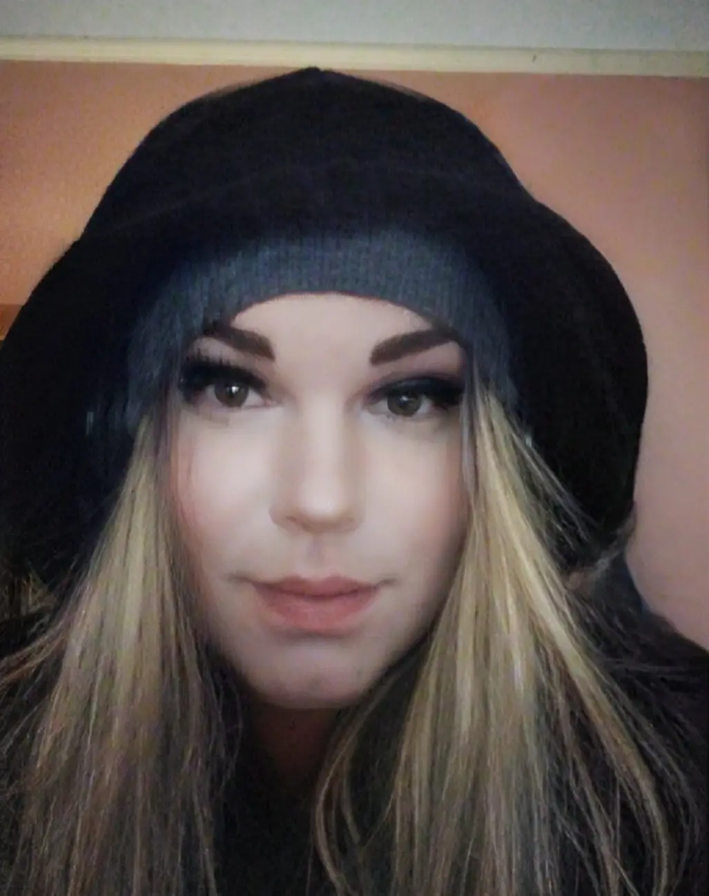 A close-up selfie of a woman with long blonde hair wearing a black beanie, looking directly at the camera with a neutral…