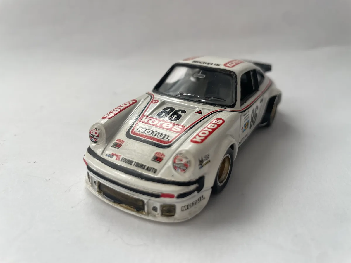 A white die-cast model of a Porsche 911 race car, number 86, with sponsor decals like 'Kores', 'Motul', and 'Michelin', sits…