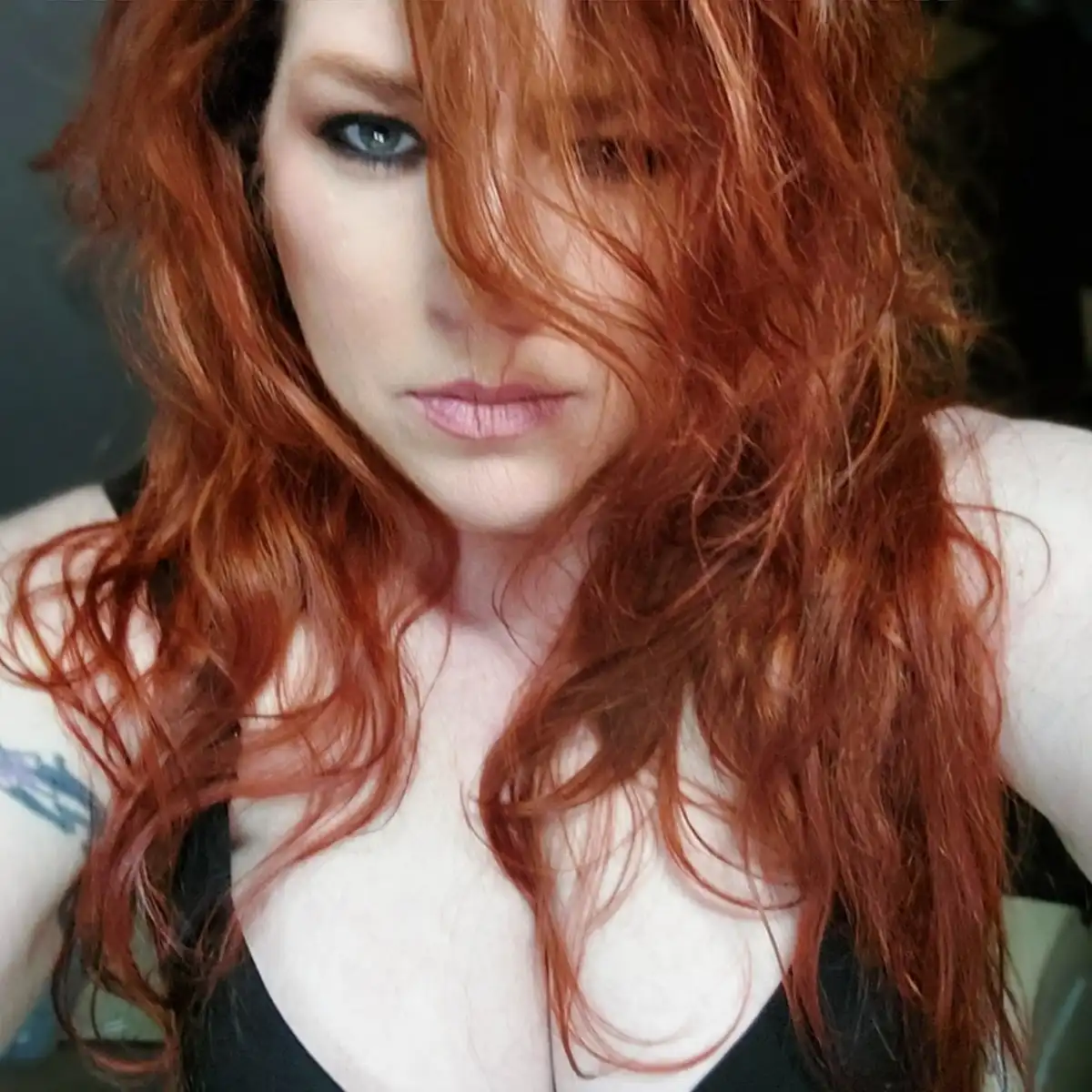 A close-up selfie of a woman with long, wavy, reddish-orange hair partially obscuring her face. She has blue eyes, light…